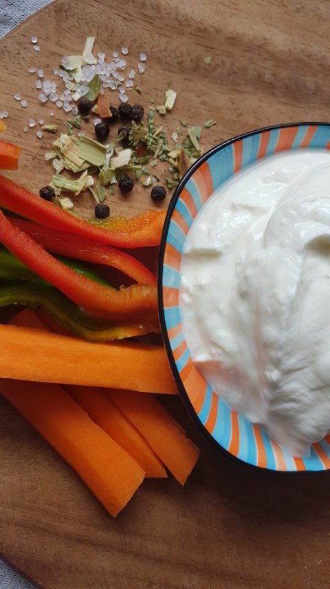 Draufsicht auf geschnittene Karotten und Paprika mit einer Schale weißen Dips und Gewürzen auf einem Holzbrett, frische und farbenfrohe Stimmung